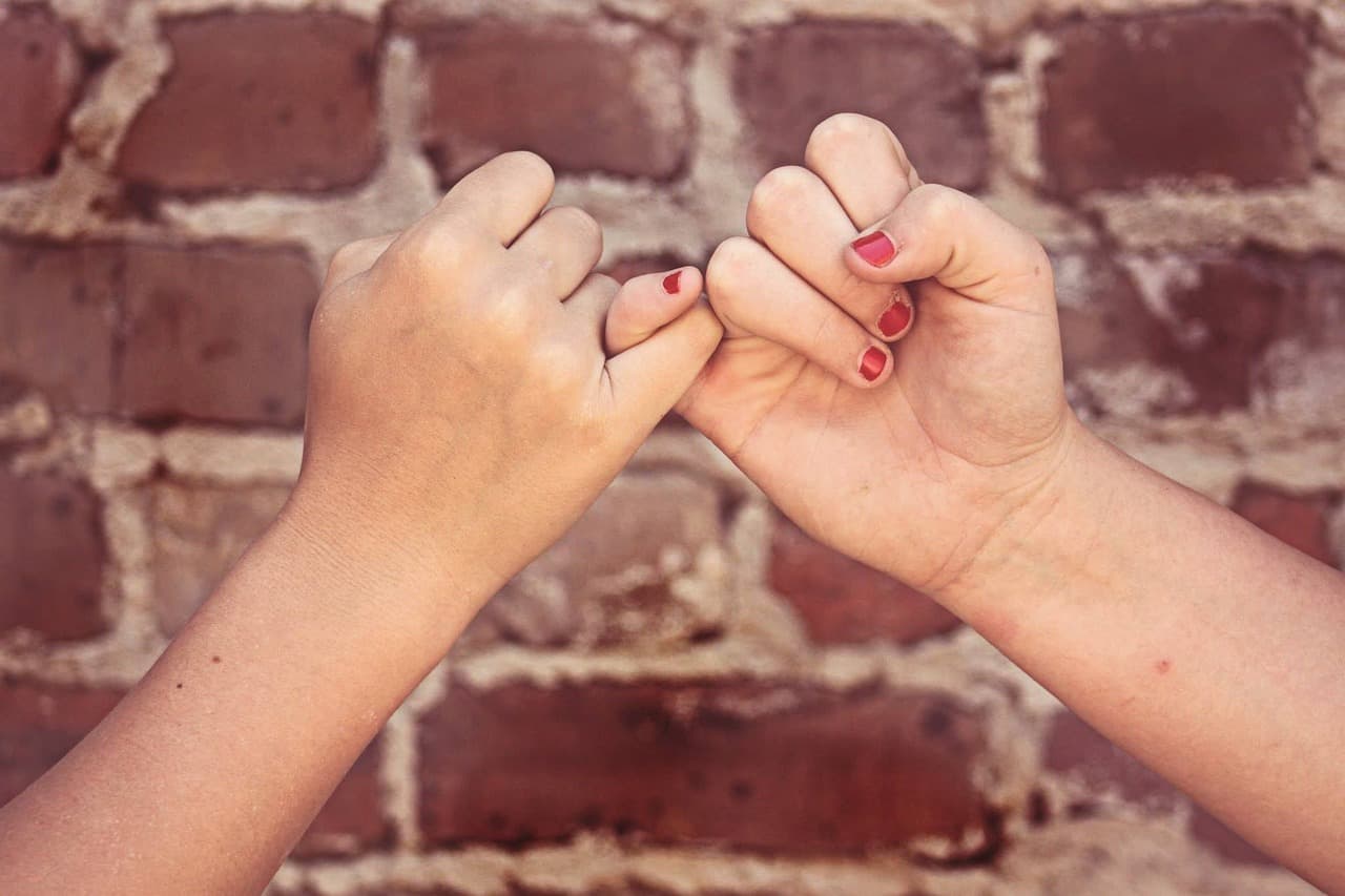 Two hands with red nail polish linking little fingers against a red brick wall