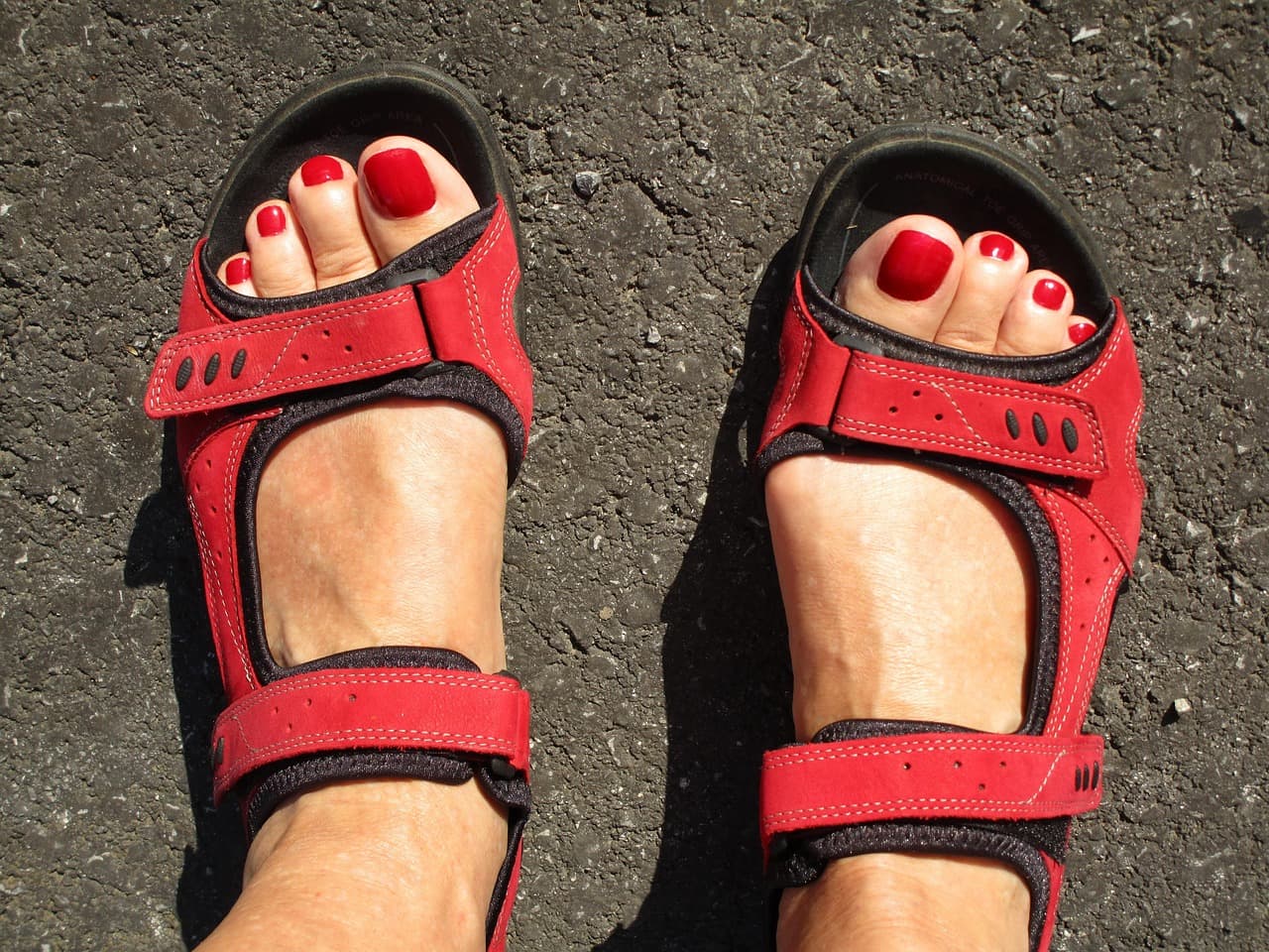 Feet with red gel toenails in red strappy sandals on dark tarmac