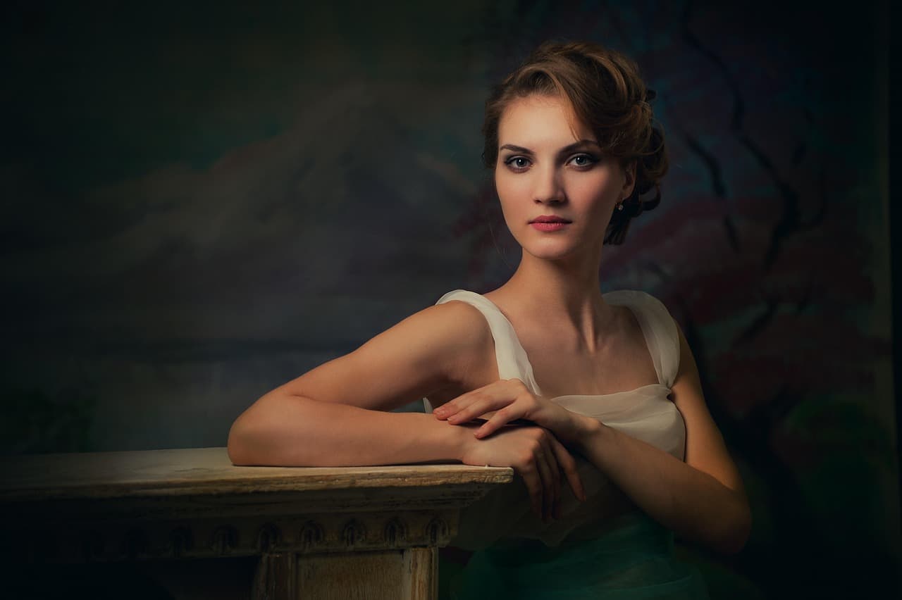 A woman in a cream dress resting her folded hands on a table, softly lit against a dark wall