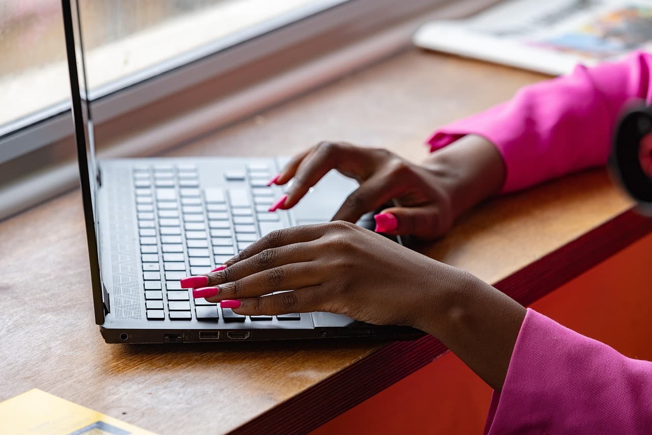 Hands with pink nails typing on a laptop at a wooden desk